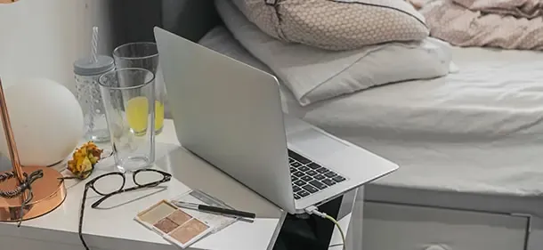 An open laptop sitting on a bedside table, next to a lamp, eyeglasses and empty drinking glasses